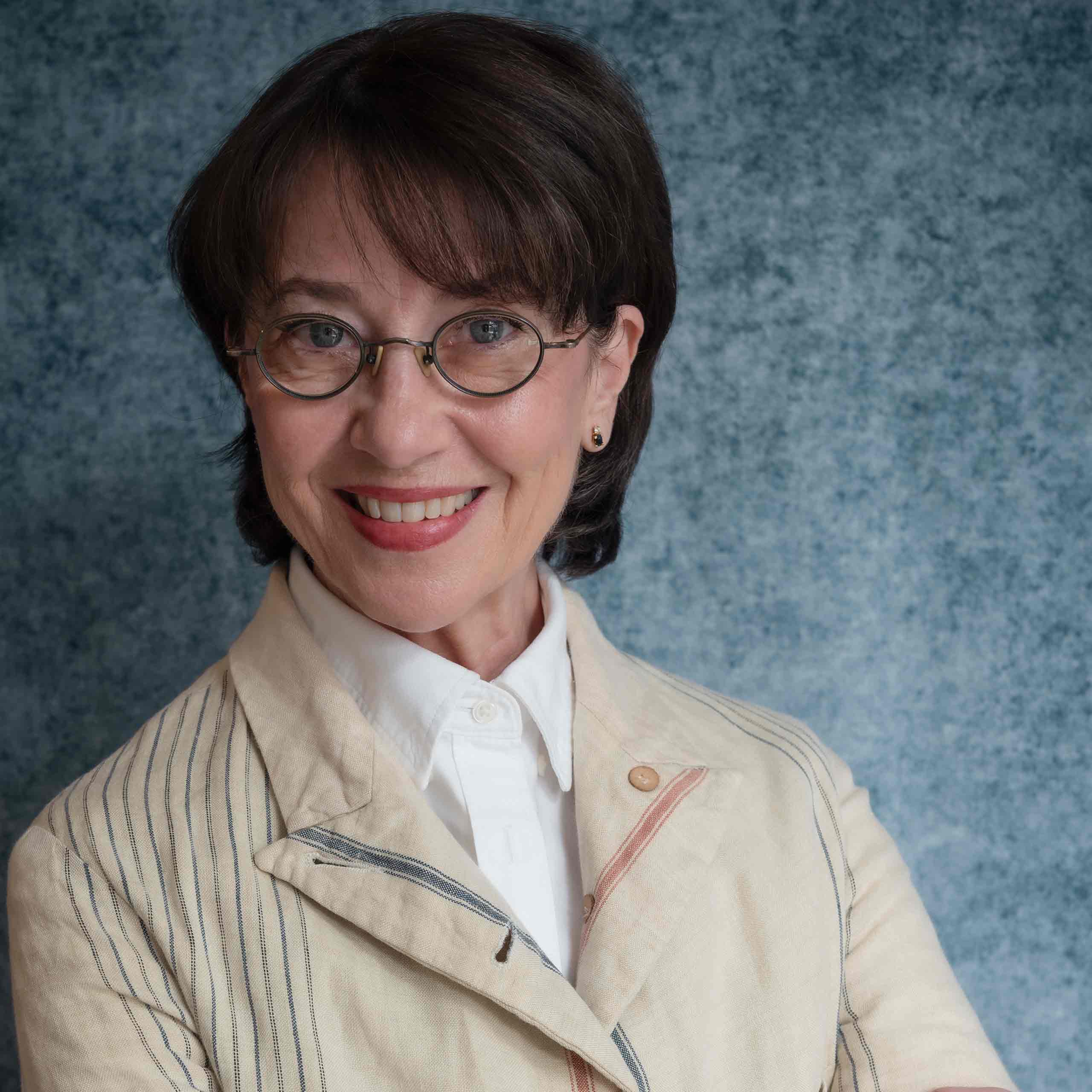 Kate Hann is a woman with medium length, dark hair. She is wearing reading glasses and a light coat and button up top.