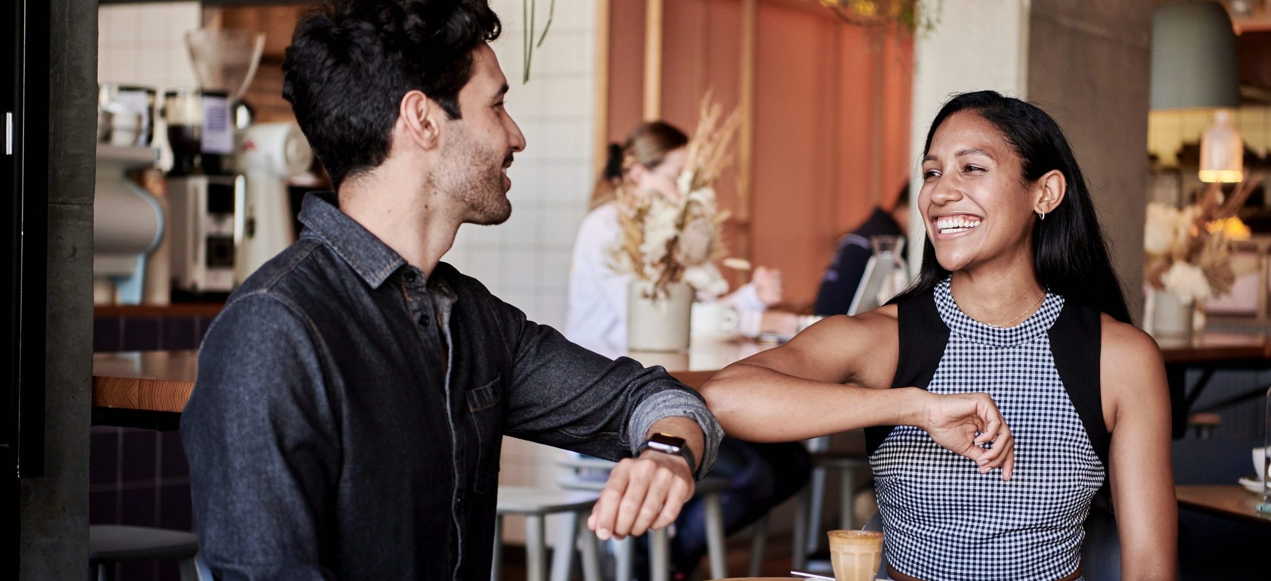 Man and woman wearing office attire are elbow bumping in a cafe. Both are smiling.