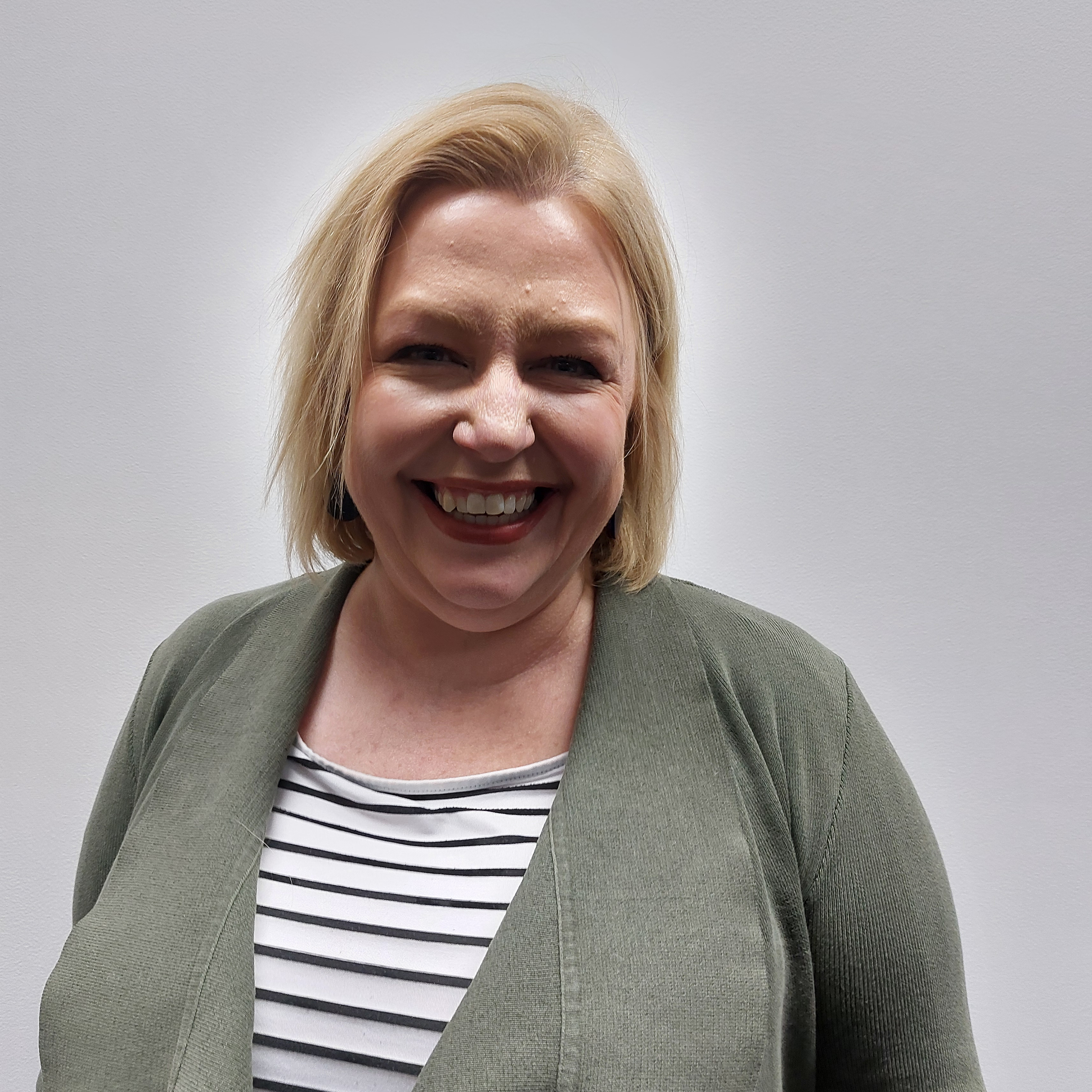 A Caucasian woman with blonde shoulder length hair and a big happy smile. She is wearing a green jacket with white stripe top.