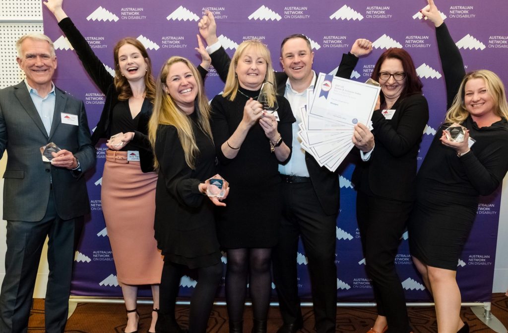 Group of seven people standing holding certificates and awards, smiling, with their hands in the air in celebration. Purple Australian Disability Network banner behind them.