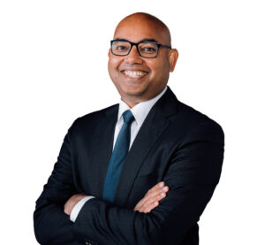 Kevin is a male wearing glasses, a collared shirt, navy blue tie and a black blazer with no hair. His arms are folded and he is smiling.