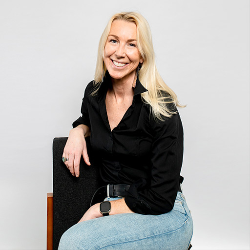 Caroline is sitting on a chair, wearing a button up shirt and jeans. She is smiling.