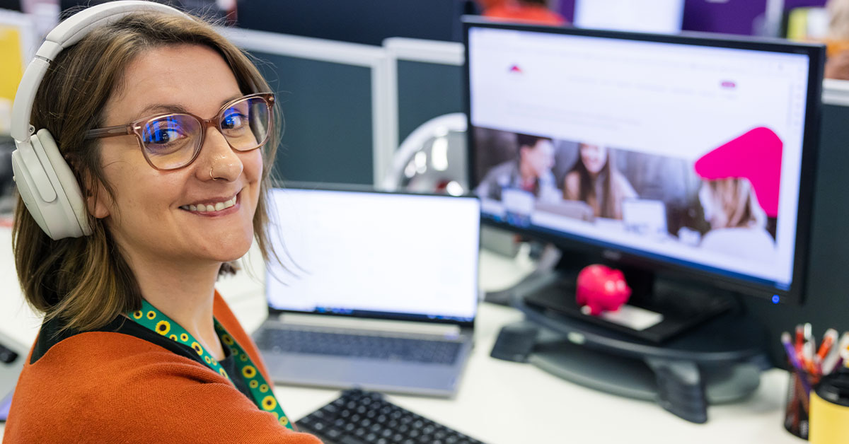 A woman with short hair smiling,. She is wearing the Hidden Disabilities Sunflower lanyard. There is a web page displaying on her monitor in the background.